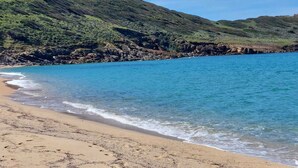 Uma praia com águas azuis cristalinas, areal arenoso e uma colina rochosa com uma torre.