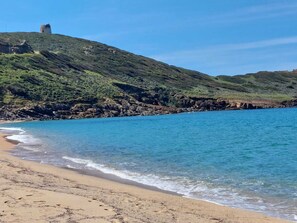 A beach with clear blue water, a sandy shore, and a rocky hill with a tower. - Dune nel Blu (Torre dei Corsari)