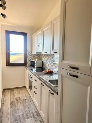A kitchen with white cabinets, a microwave, a sink, and a window with a view. - Dune nel Blu (Torre dei Corsari)