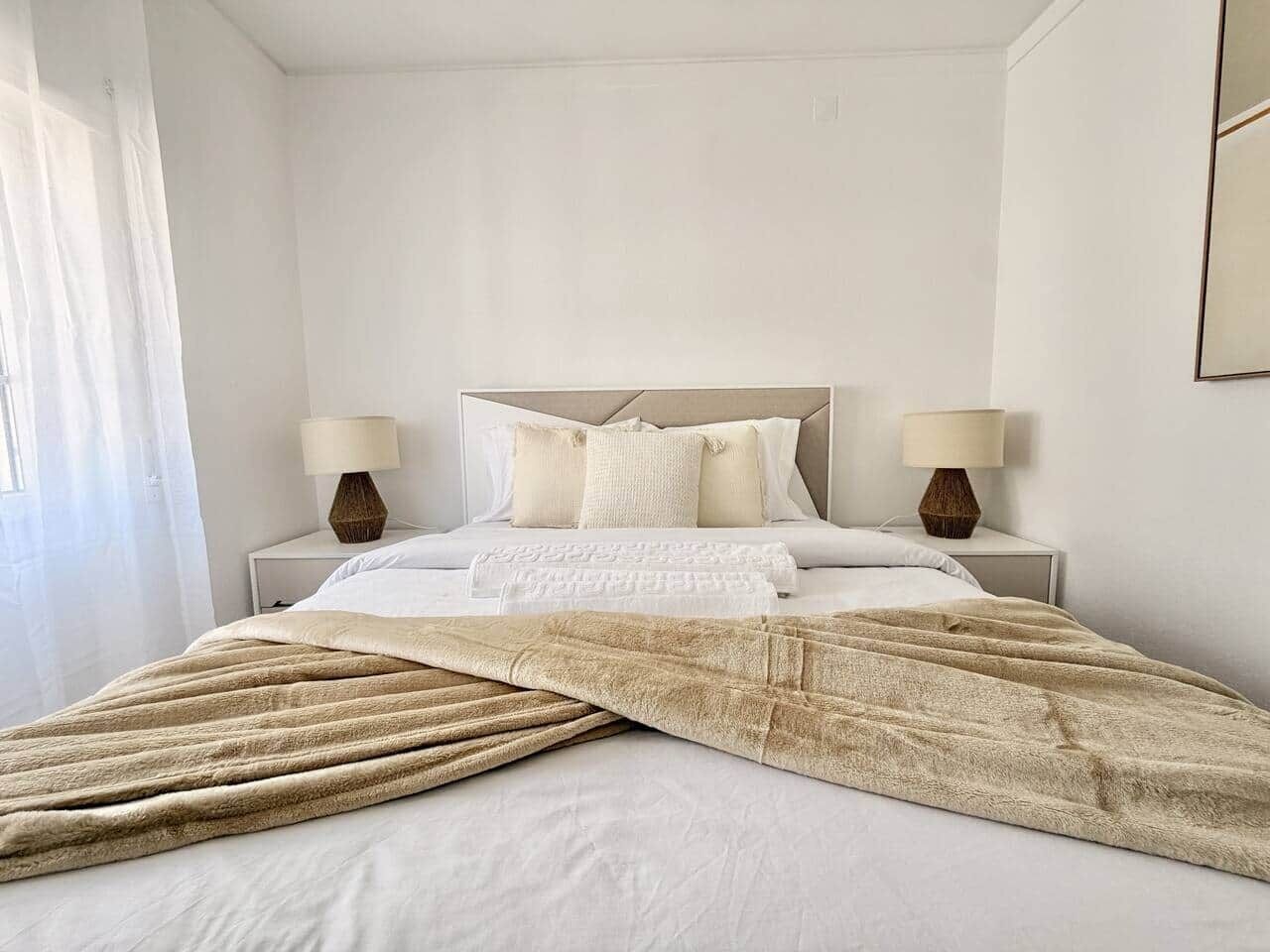 Ein ordentlich bezogenes Bett mit einer beige farbenen Decke, zwei Nachttischlampen und einem Kopfteil.