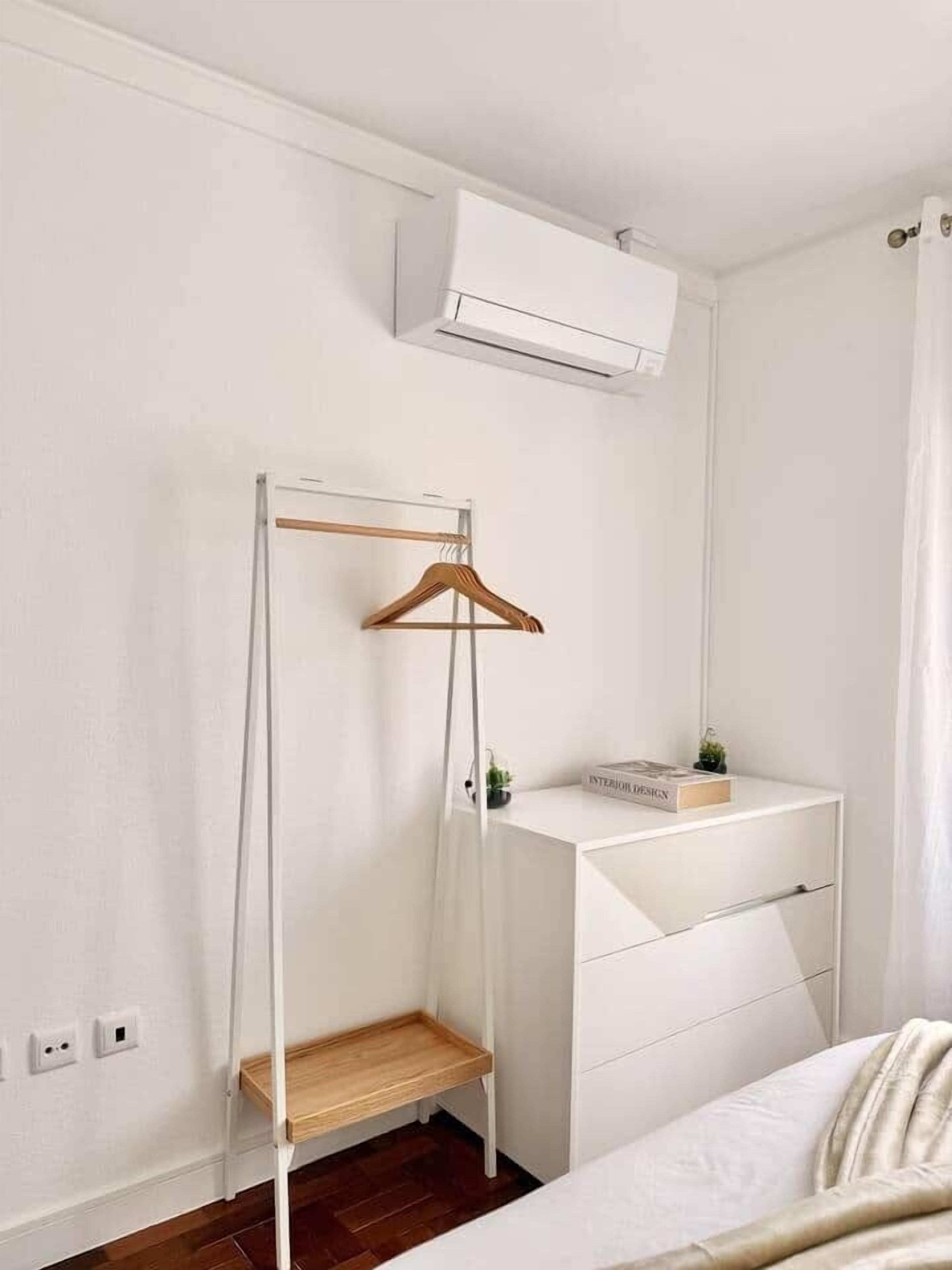 Un dormitorio con una cama blanca, un estante de madera, un perchero y una mesita de noche con un libro y una planta.