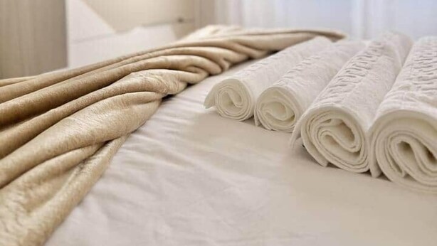 Una cama bien hecha con toallas enrolladas y una manta color beige en una habitación luminosa con ventana.