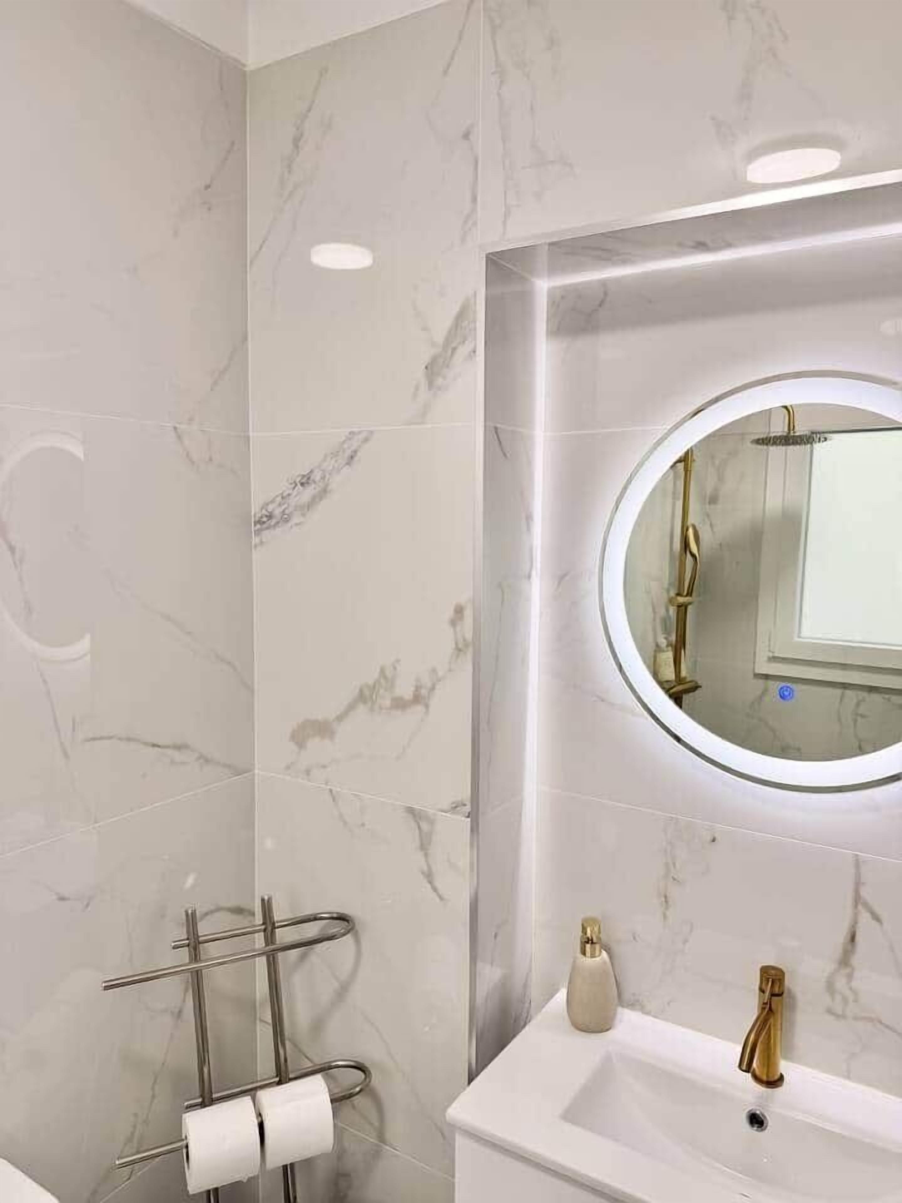 A bathroom with marble walls, a round mirror, a white sink with a gold faucet, and a towel rack with white towels.