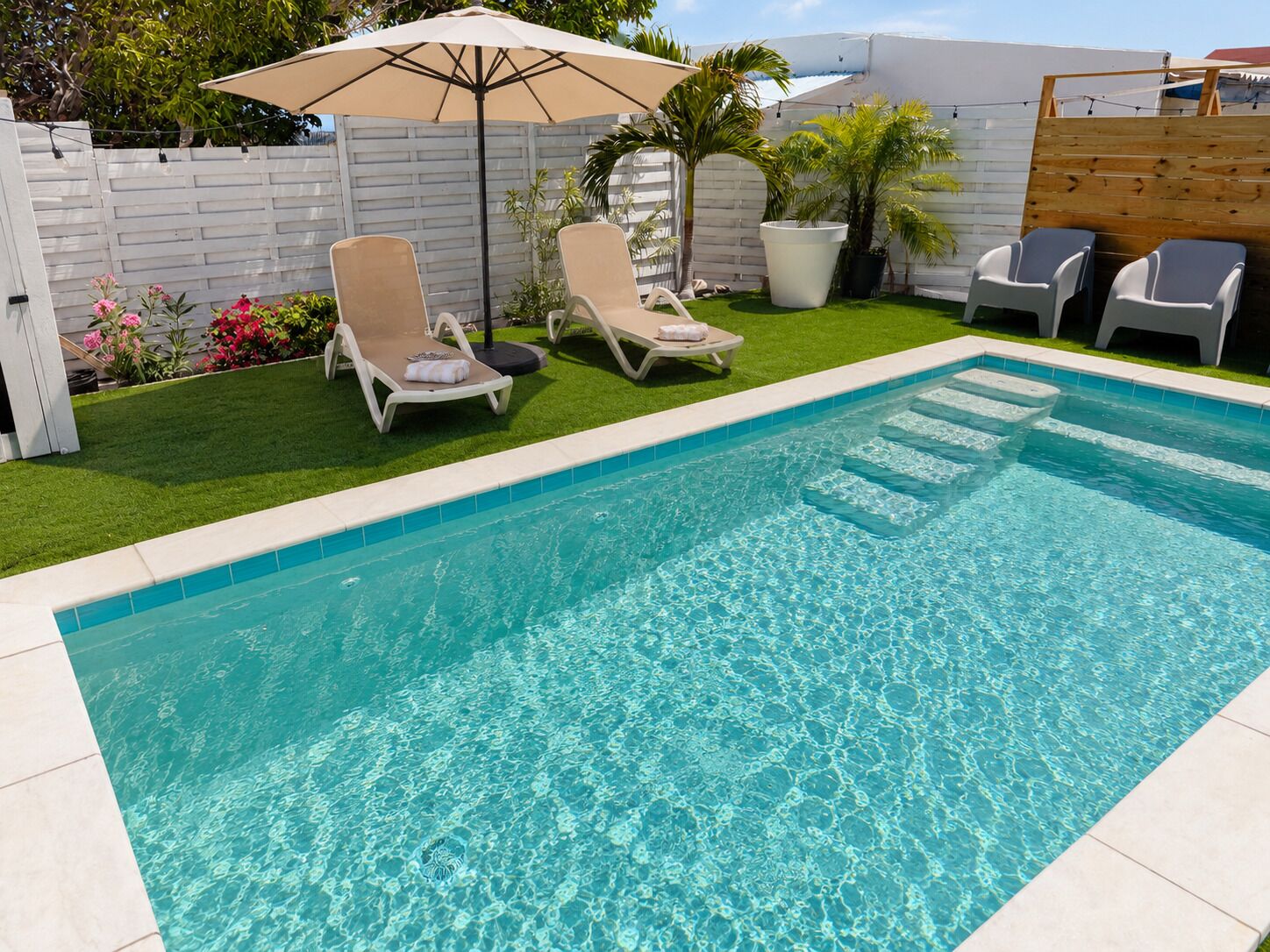 Une piscine rectangulaire à l’eau d’un bleu limpide, entourée d’une terrasse carrelée de blanc. On y trouve deux chaises longues avec des serviettes, un grand parasol et, au fond, une clôture en bois.