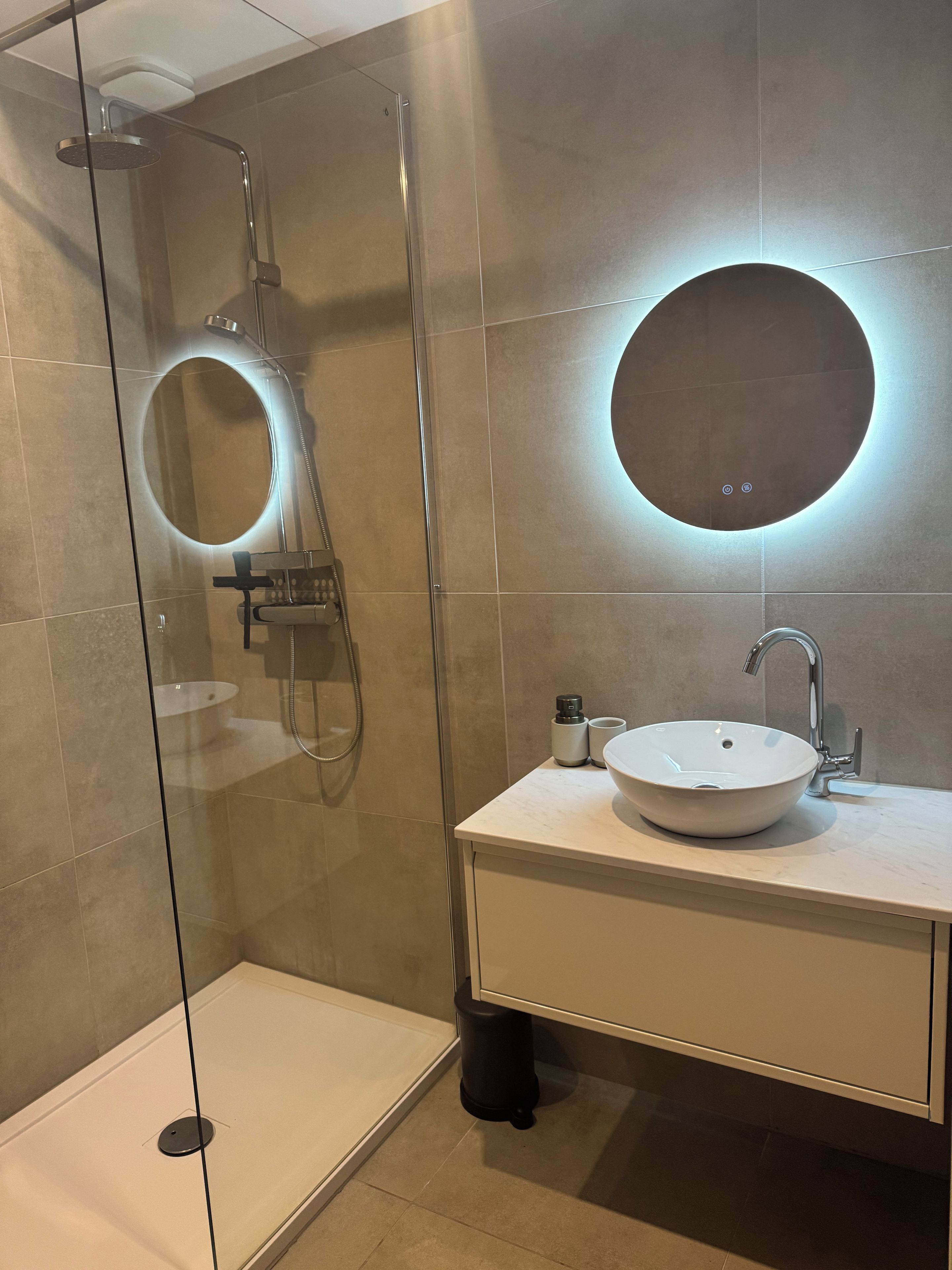 Ein modernes Badezimmer mit einer gläsernen Dusche, einem runden Spiegel und einem weißen Waschbecken mit Armatur.
