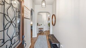 Un couloir avec une porte en bois, un portail en métal, un luminaire suspendu, un tapis, un banc et une citation encadrée accrochée au mur.