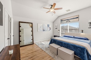 A bedroom with a bed, a ceiling fan, a mirror, artwork on the wall, and a window with blinds.
