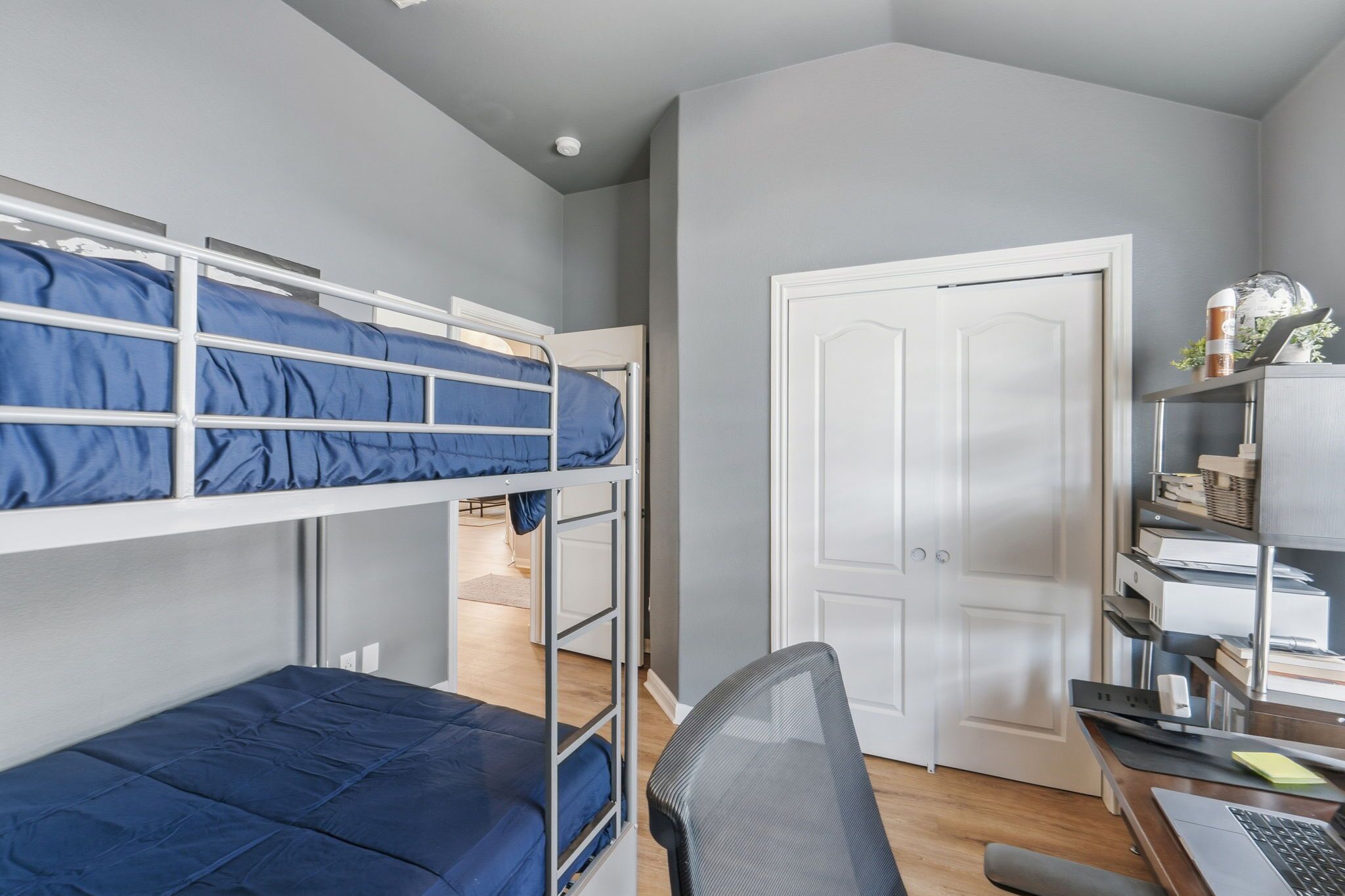 A bunk bed room with a desk and chair, a white door, and a shelf with books and a lamp.