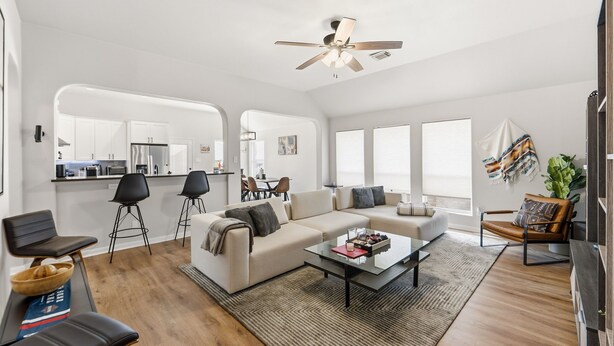 Un salon moderne avec un canapé, une table basse et des fauteuils, une cuisine en arrière-plan et un ventilateur de plafond.