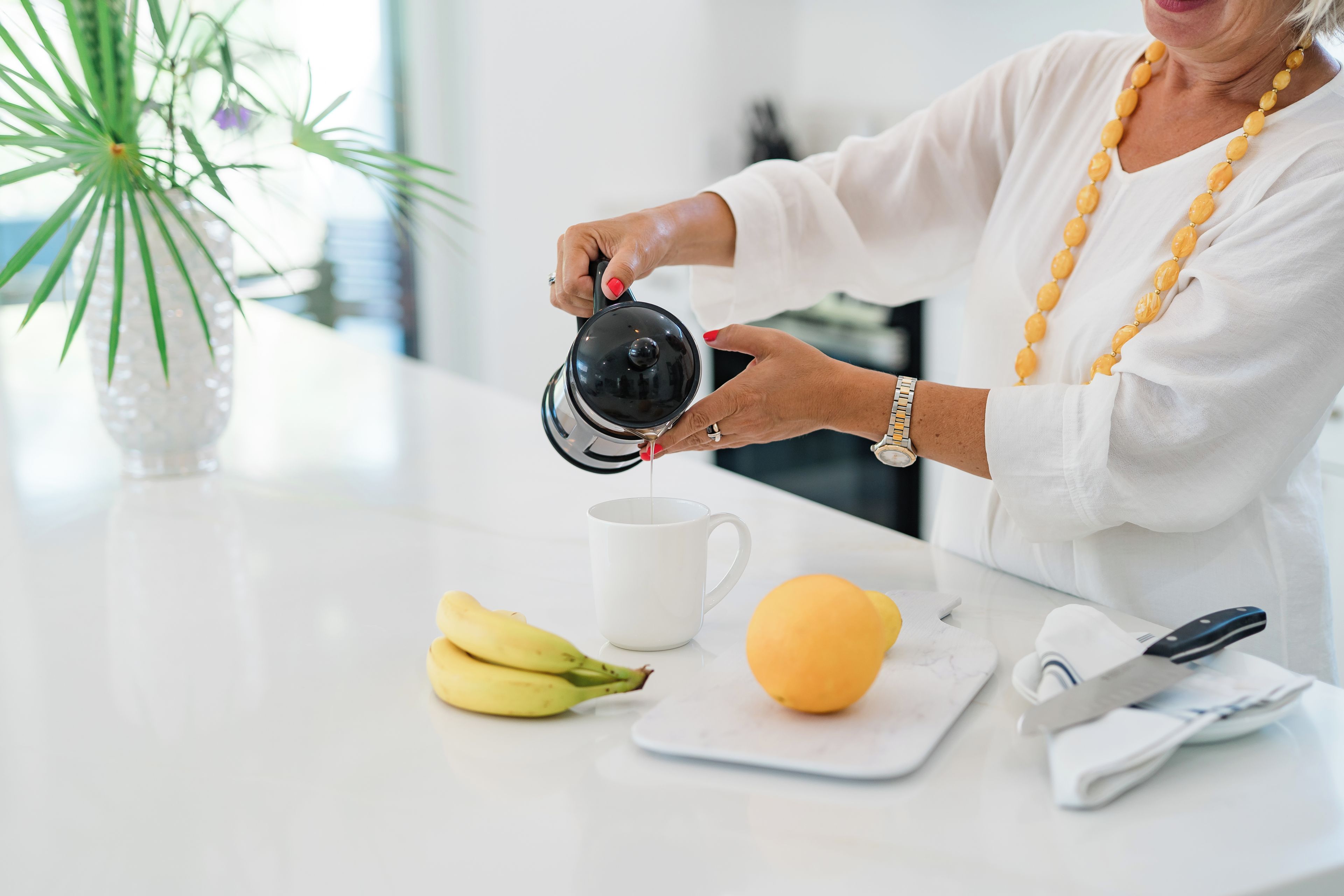 Una persona vertiendo líquido de una tetera en una taza blanca, con plátanos y un limón sobre la encimera.