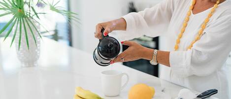 Una persona vertiendo líquido de una tetera en una taza blanca, con plátanos y un limón sobre la encimera.