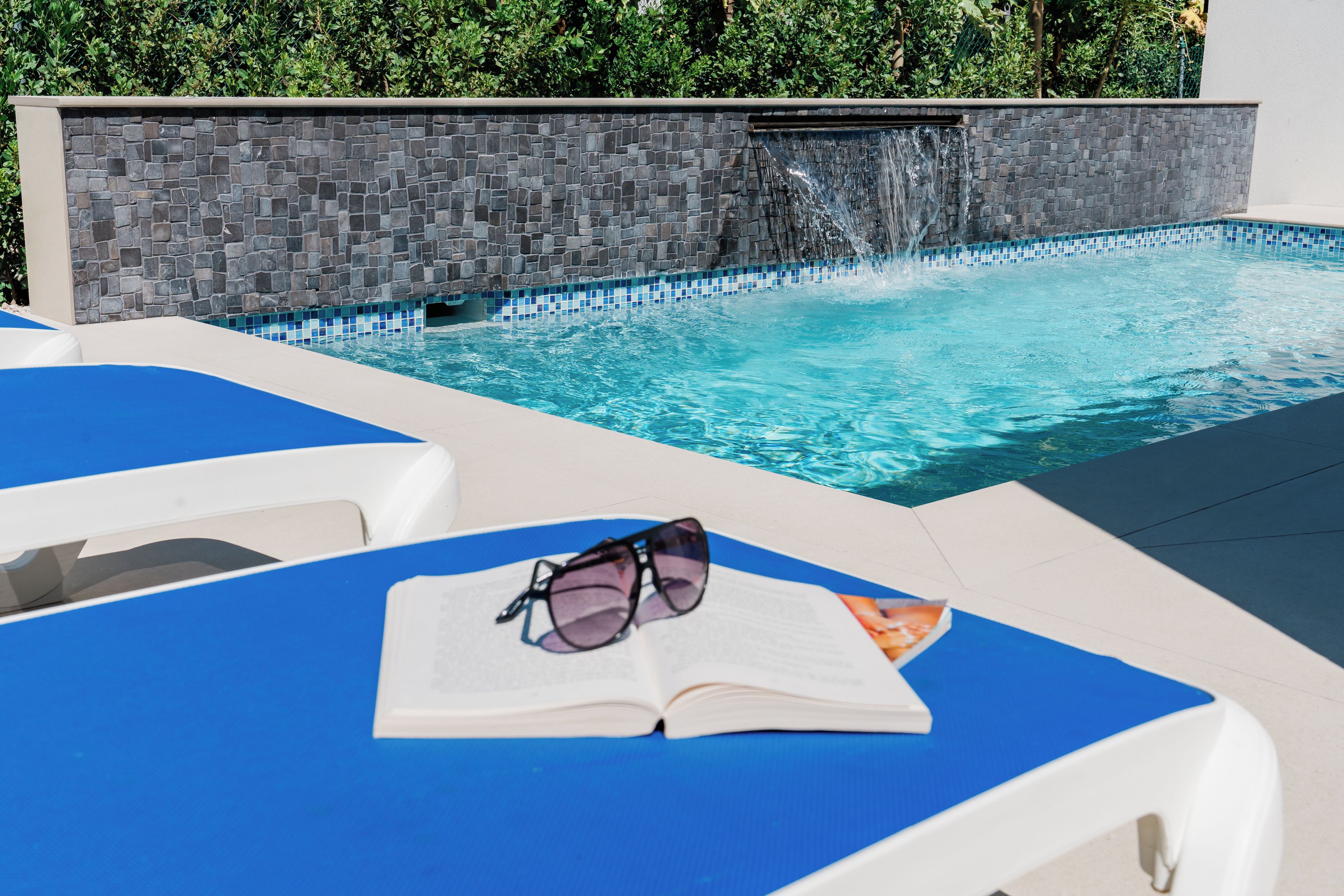 A swimming pool with a stone wall and a water feature. There are lounge chairs and a book with sunglasses on one of them.