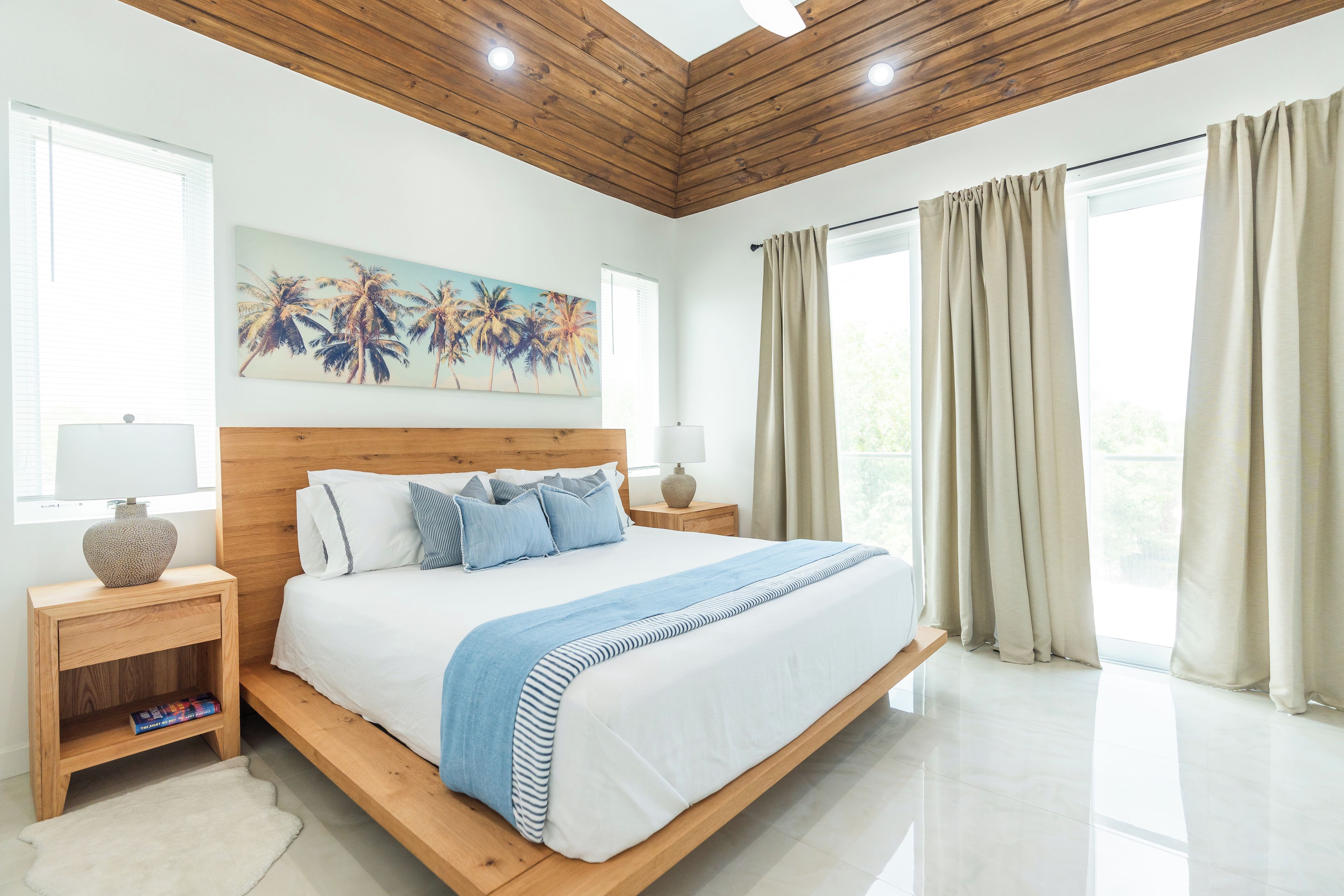 A bedroom with a wooden bed frame, white bedding, a blue-striped throw, and a painting of palm trees.