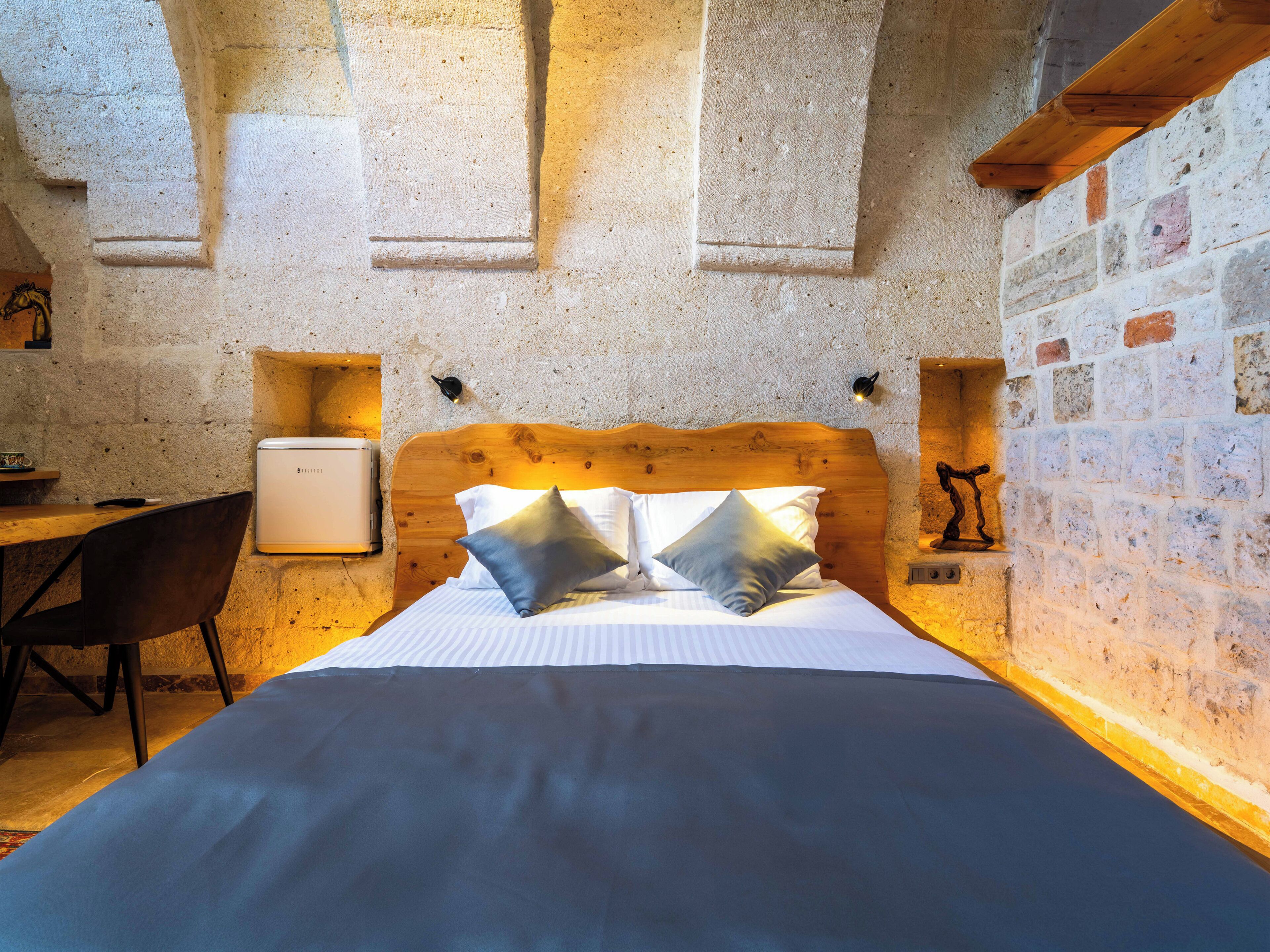 A bedroom with a wooden headboard, a bed with pillows, a desk, and a small refrigerator.