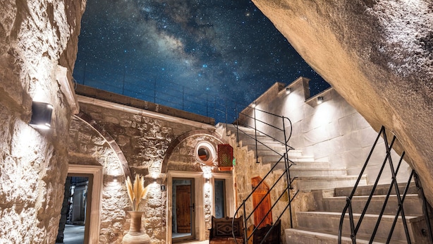 Un intérieur aux murs de pierre, avec un escalier menant à un deuxième étage, une porte en bois et un ciel étoilé visible à travers une lucarne.