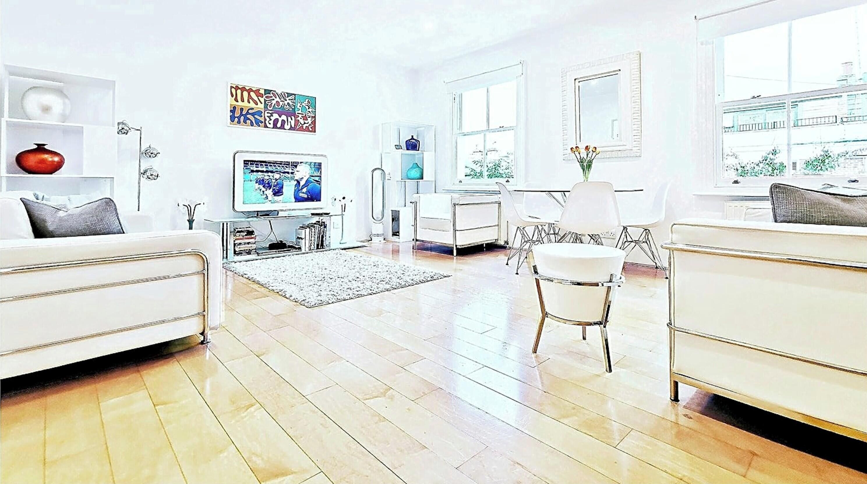 A modern living room with a flat-screen TV, white sofas, a dining area with chairs, and a kitchen area.
