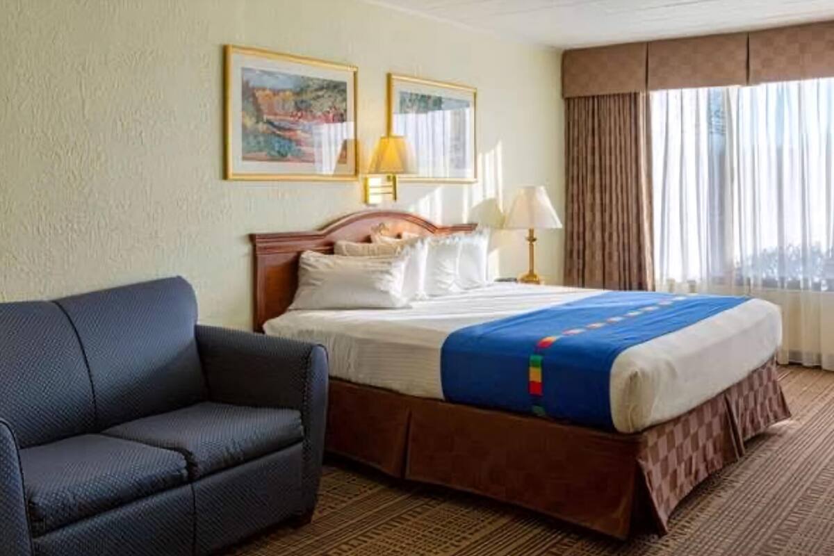 A hotel room with a bed, a sofa, two framed pictures on the wall, a window with curtains, and a lamp.