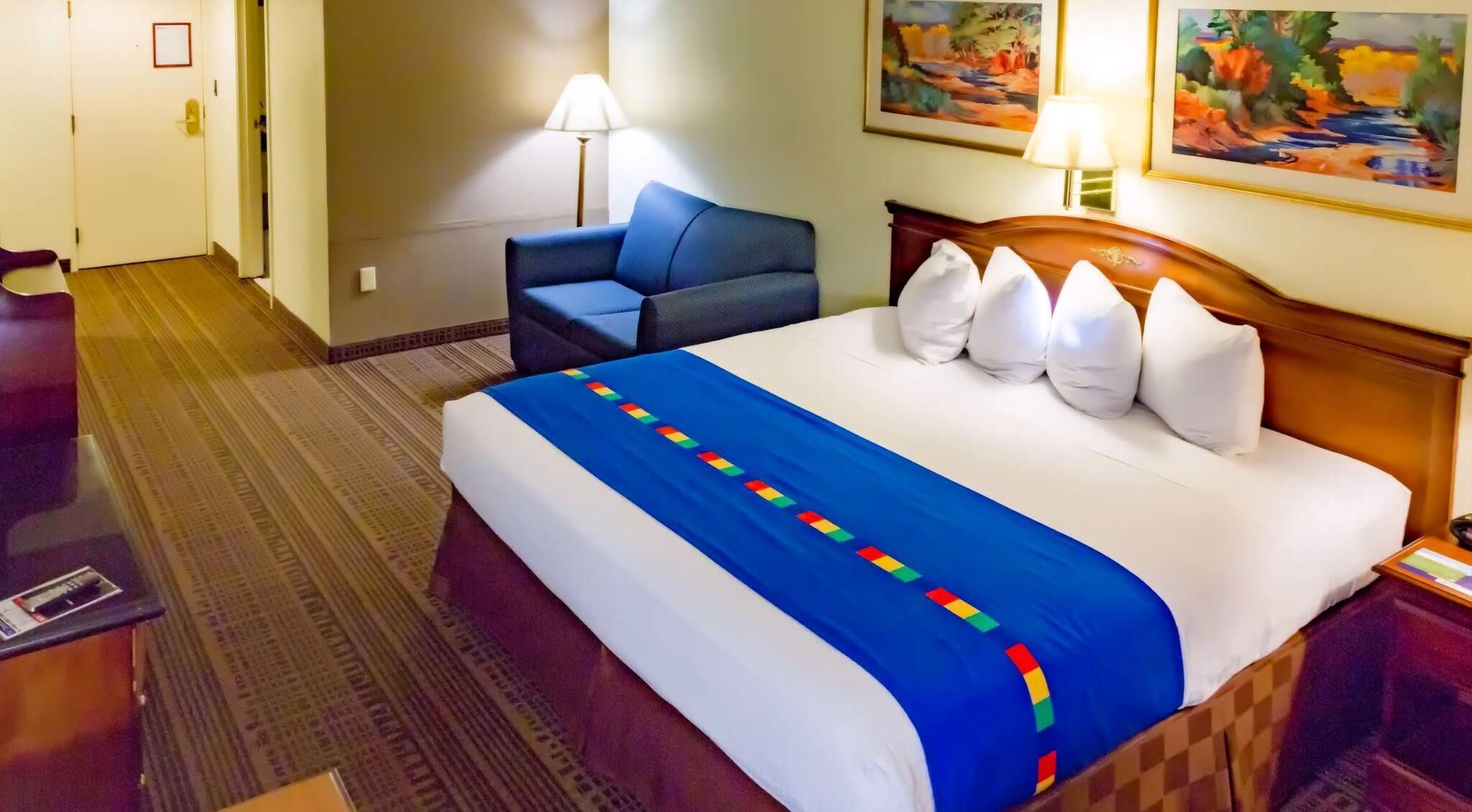 A hotel room with a bed, a chair, a desk, and two paintings on the wall.
