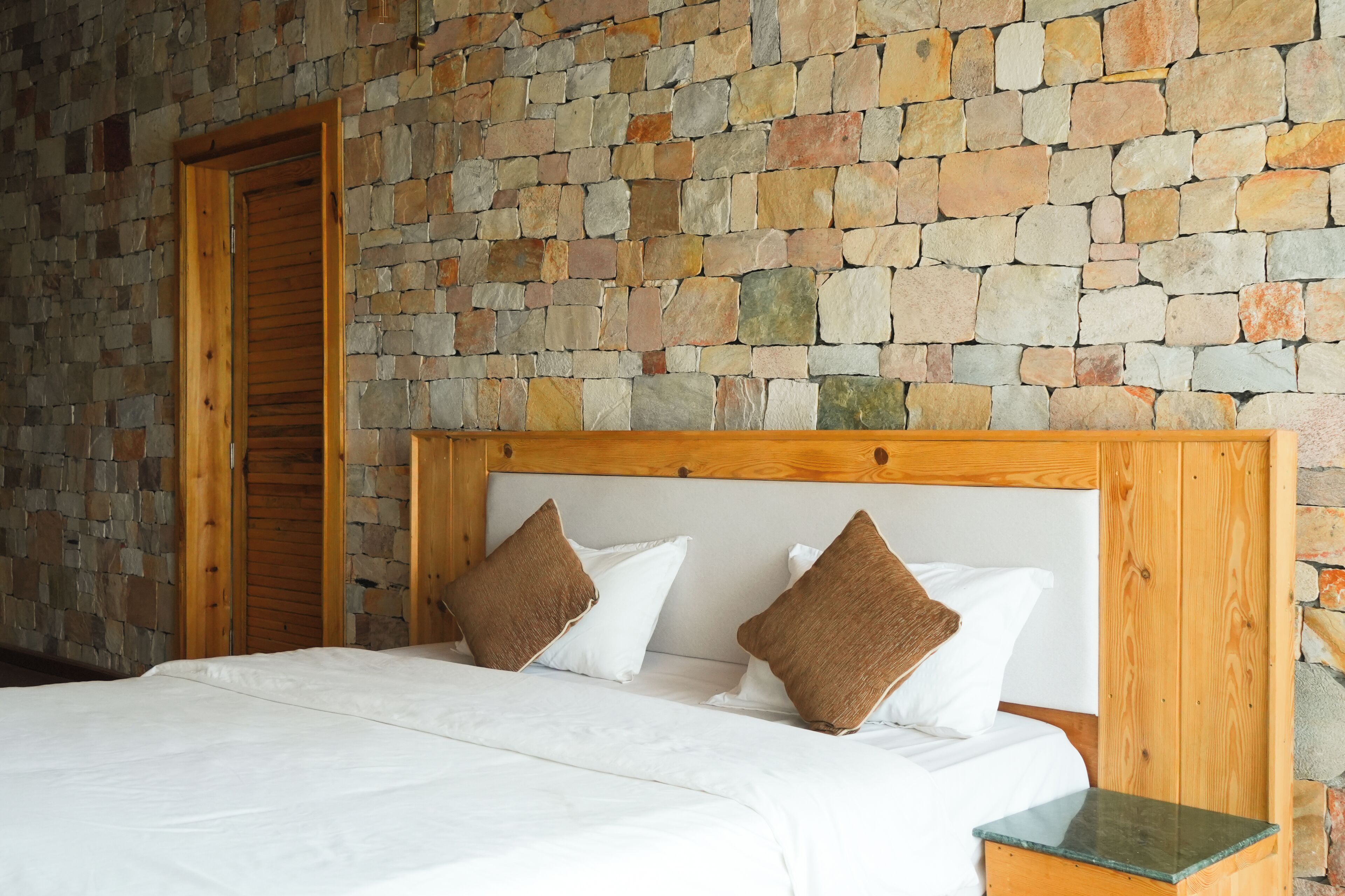 Un lit avec une tête de lit en bois, une literie blanche et des oreillers marron, dans une pièce dont un des murs est en pierre.