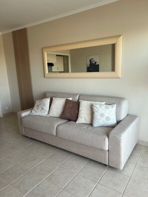 A living room with a sofa, a large mirror, and a tiled floor. - Quiet studio with balcony and sea view, downtown, 10 minutes walk from the beaches and the palace (Cannes)