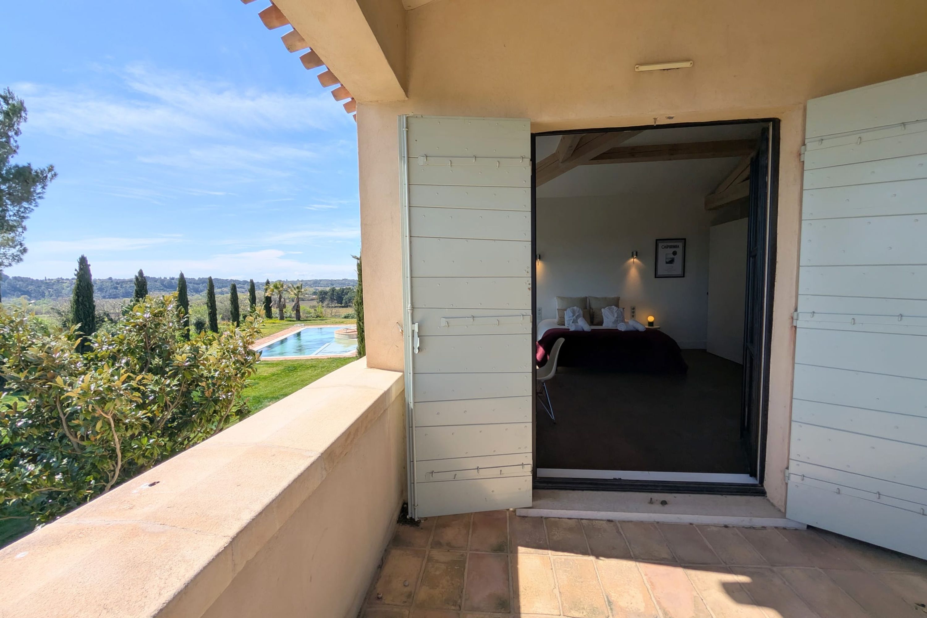 An open door to a bedroom with a bed and a nightstand, a view of a swimming pool, and a clear sky.
