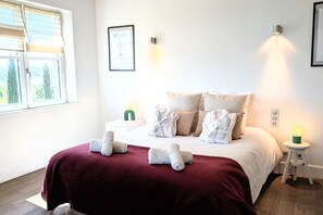 A neatly made bed with a red bedspread, a window with blinds, and a wall-mounted light fixture. - Wine Estate 20m Pool and Cinema (Pezenas)