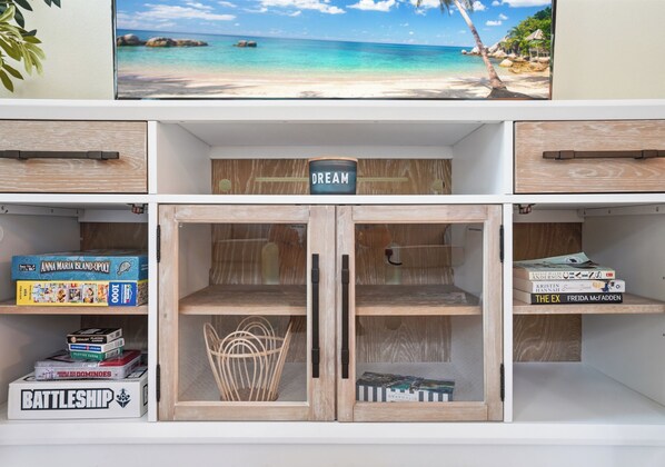 A room with a television displaying a beach scene, wooden cabinets, and shelves with books and board games. - Stylish 3BR Retreat Close to AMI Beaches (bradenton)