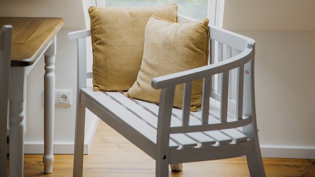Una silla de madera blanca con cojín, ubicada debajo de un ventanal con vistas al verdor exterior.