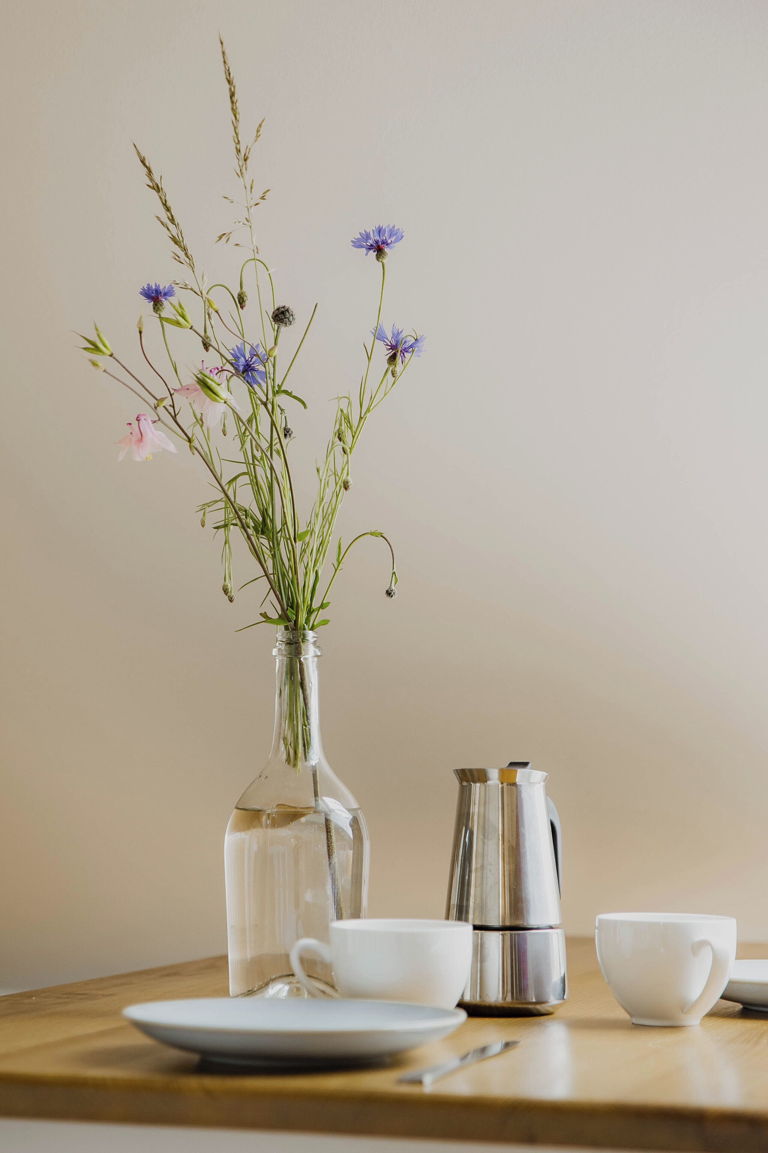 Un florero de vidrio transparente con flores sobre una mesa de madera, junto con una cafetera de plata y dos tazas blancas.
