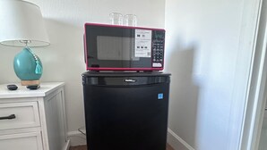 A compact black refrigerator with a microwave on top, a white nightstand with a teal lamp, and a wooden floor. - Hotel Style 1-bedroom in lovely Orlando with AC (Orlando)