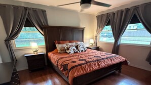 A bedroom with a bed, wooden floor, ceiling fan, nightstands, and curtains.