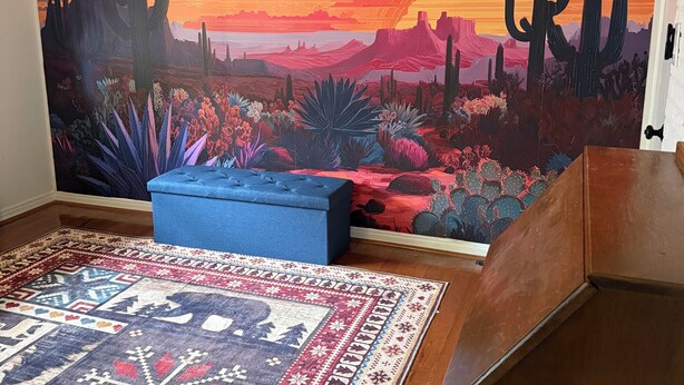 A room with a large mural of a desert sunset, a blue cushioned bench, and a patterned rug.
