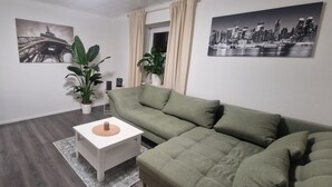 A living room with a green sofa, a white side table, a potted plant, and two wall-mounted pictures. - Vacation apartment Lichtblick in a quiet location / free parking and WLAN (Sulingen)