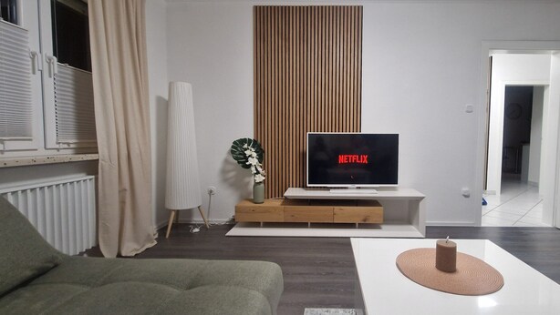 A modern living room with a flat-screen TV, a green sofa, a white coffee table, and a wooden accent wall. - Vacation apartment Lichtblick in a quiet location / free parking and WLAN (Sulingen)