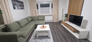 Una sala de estar con un sofá modular, una mesa de centro, un televisor de pantalla plana sobre un mueble, un radiador y una ventana con cortinas.