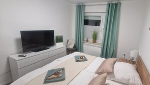 A bedroom with a bed, a TV on a stand, a small table with a lamp, and a window with curtains. - Vacation apartment Lichtblick in a quiet location / free parking and WLAN (Sulingen)