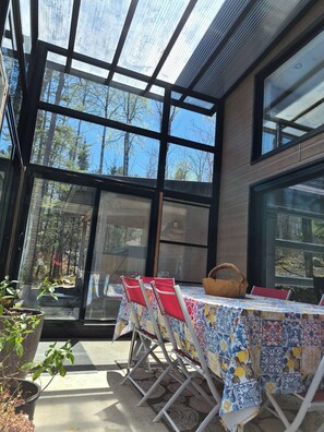 A sunlit outdoor dining area with a glass roof, red chairs, and a table with a patterned tablecloth.