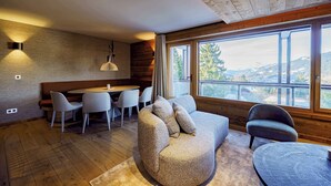 Een knus vertrek met een houten plafond, een ronde eettafel met stoelen, een bank met kussens en grote ramen met uitzicht op de bergen.