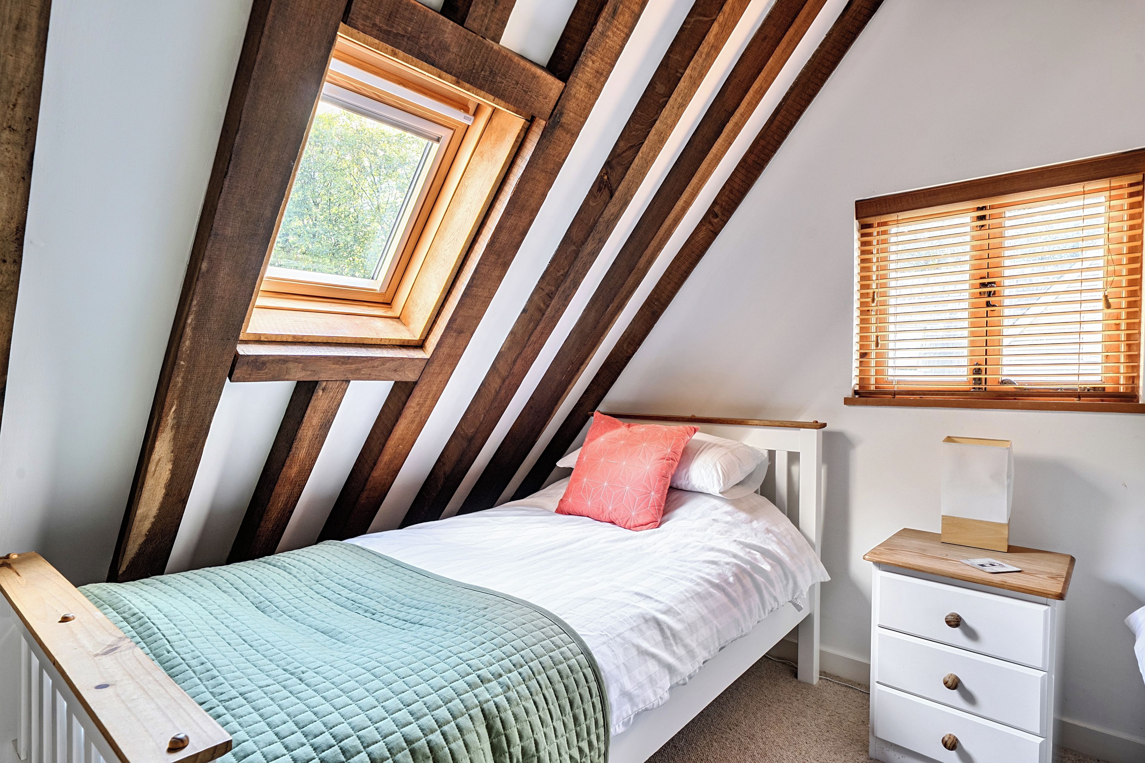 Ein Schlafzimmer mit schrägen Wänden, einem Bett mit gesteppter Bettdecke, einem Nachttisch mit Lampe und einem Fenster mit Holzjalousien.