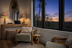 Ein gemütliches Zimmer mit einem großen Fenster, einem Schreibtisch mit einem Laptop, zwei Lampen und einem Sessel mit einem Glas Wein.