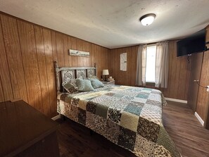 Un dormitorio con pared de paneles de madera, una cama con colcha, una mesita de noche, una l ámpara, un televisor montado en la pared y una ventana con cortinas.