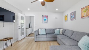 Ein modernes Wohnzimmer mit grauem Ecksofa, einem Holztisch, einem Deckenventilator und Wandbildern.