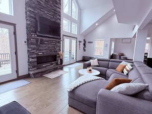 A spacious living room with a stone fireplace, a grey sectional sofa, and a flat-screen TV. - Luxury 5BR Chalet with Lake, Pool, Ski, Game Room, 2 Decks and Sleeps 14 (Lackawaxen)