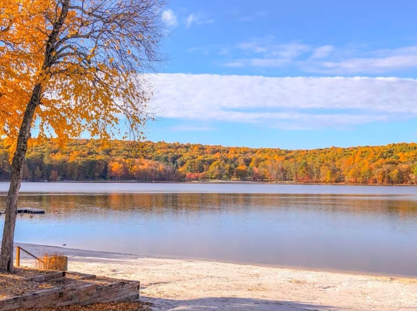 A lake with autumn foliage, a sandy beach, and a wooden dock.