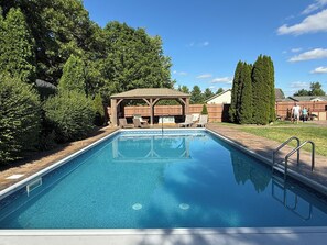 A rectangular swimming pool with a wooden deck and lounge chairs, surrounded by trees and a covered pavilion.