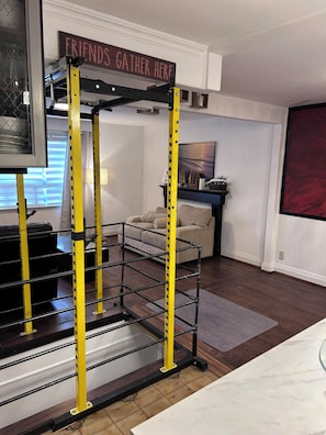 A home gym with a yellow metal frame, a couch, a painting, and a sign that says 'FRIENDS GATHER HERE'.