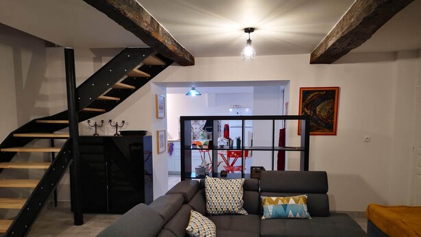 A modern living room with a dark-colored sofa, patterned cushions, a wooden staircase, and a kitchen area in the background. - Port House (Bourg)