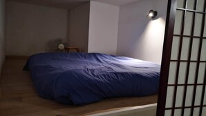 A room with a bed, a small table, a fan, and a wall-mounted light fixture. - Port House (Bourg)