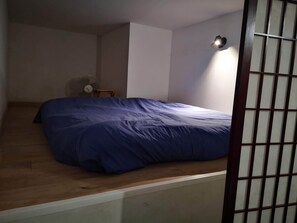 Una habitación con una cama, una mesita, un ventilador y una lámpara fijada a la pared.