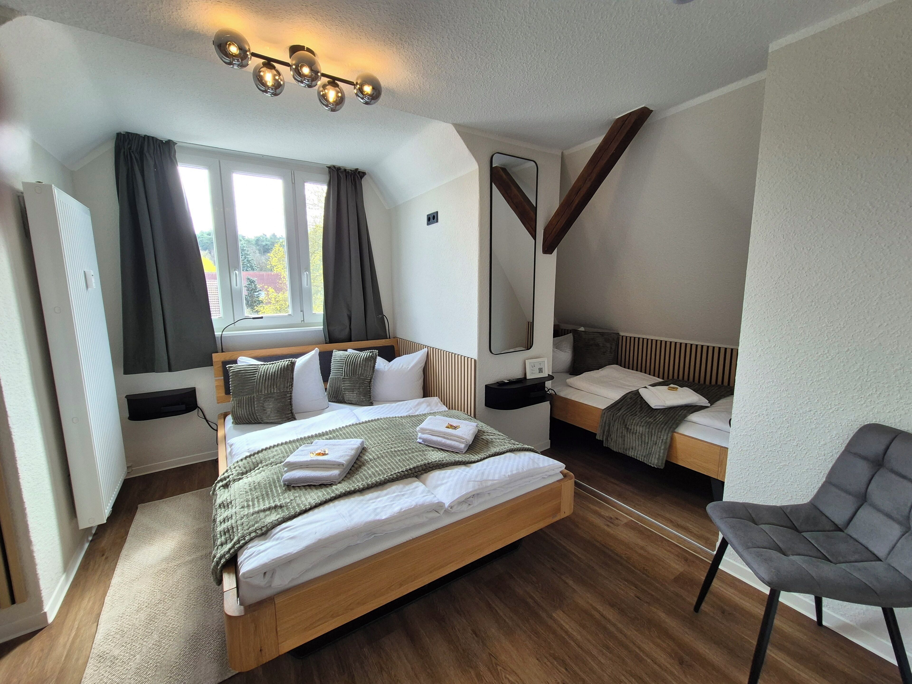 Ein modernes Hotelzimmer mit einem großen Bett, einem kleineren Bett, einem Sessel, einem Spiegel und einem Fenster mit Vorhängen.